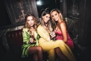 Three women in a bar, seated and smiling at the camera; one in a green dress holding a cocktail, another in a yellow outfit holding a glass of champagne, and the third in a pink dress holding a drink.