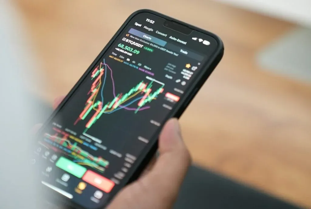 A person holding a smartphone displaying a cryptocurrency trading app, showing a candlestick chart and various trading data.