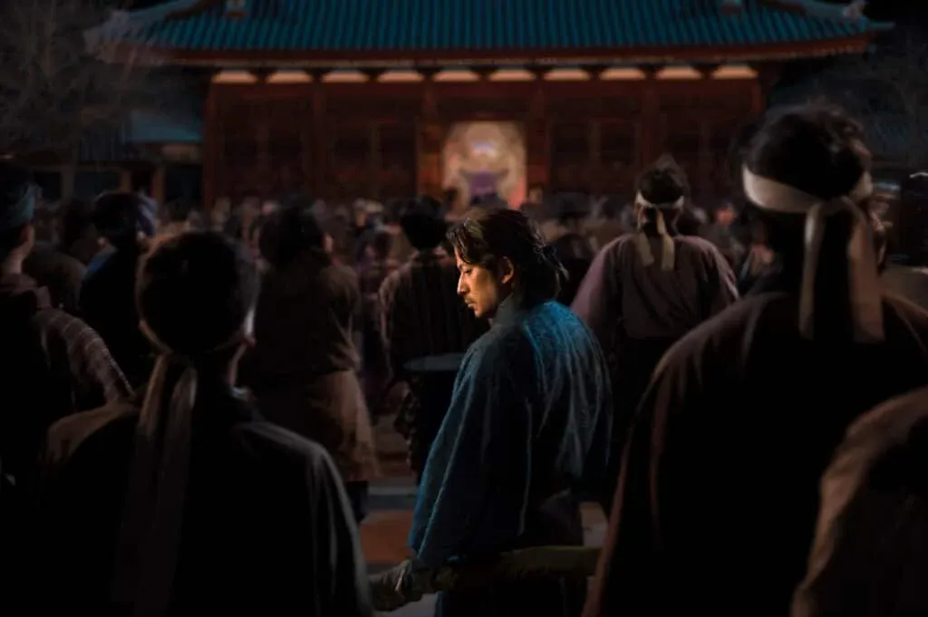 A scene depicting a large group of people gathered in front of a traditional building with a blue-tiled roof. The focus is on a man in a blue robe standing in the foreground, looking down thoughtfully, surrounded by others in period attire.