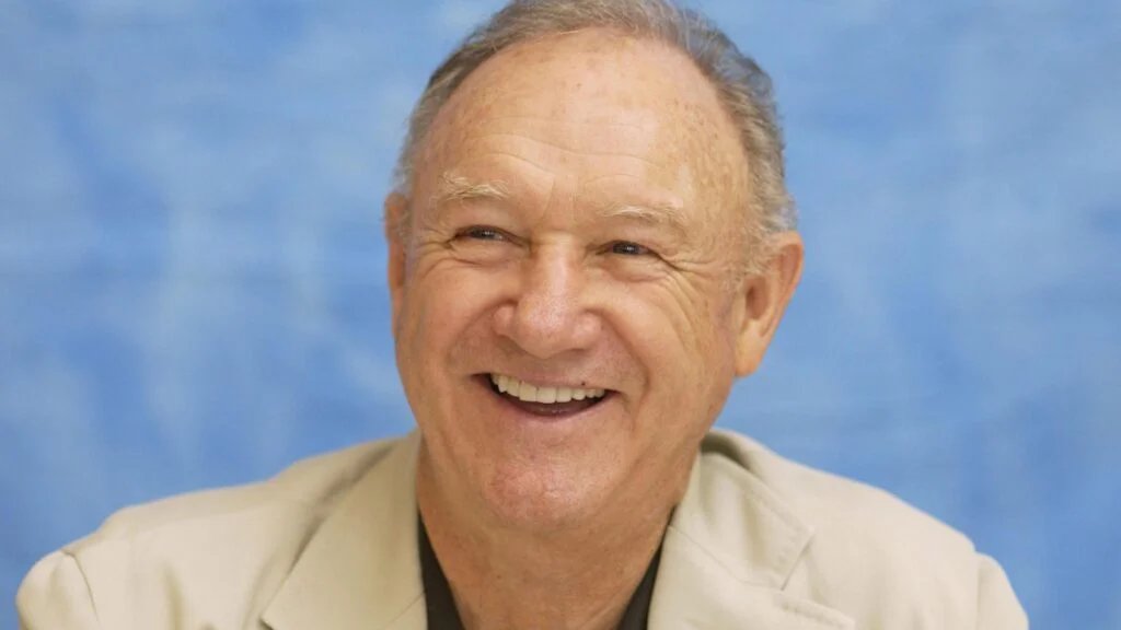 A smiling elderly man wearing a light-colored suit with a blue background.