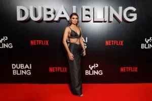 A woman stands on a red carpet in front of a wall with the words 'Dubai Bling' and 'Netflix' displayed in large letters. The setting appears to be a formal event or premiere.