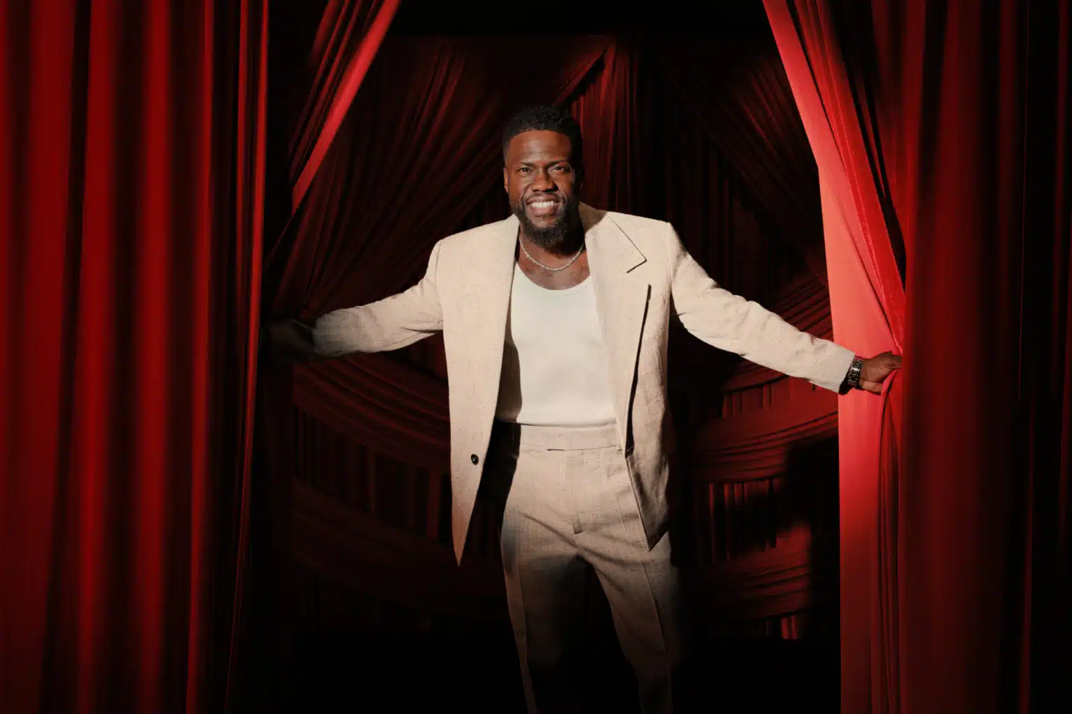 A man in a light beige suit and a white shirt stands smiling, holding apart red theater curtains.