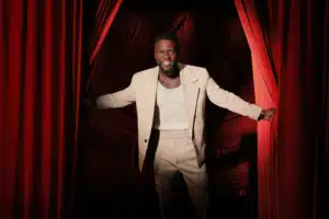A man in a light beige suit and a white shirt stands smiling, holding apart red theater curtains.