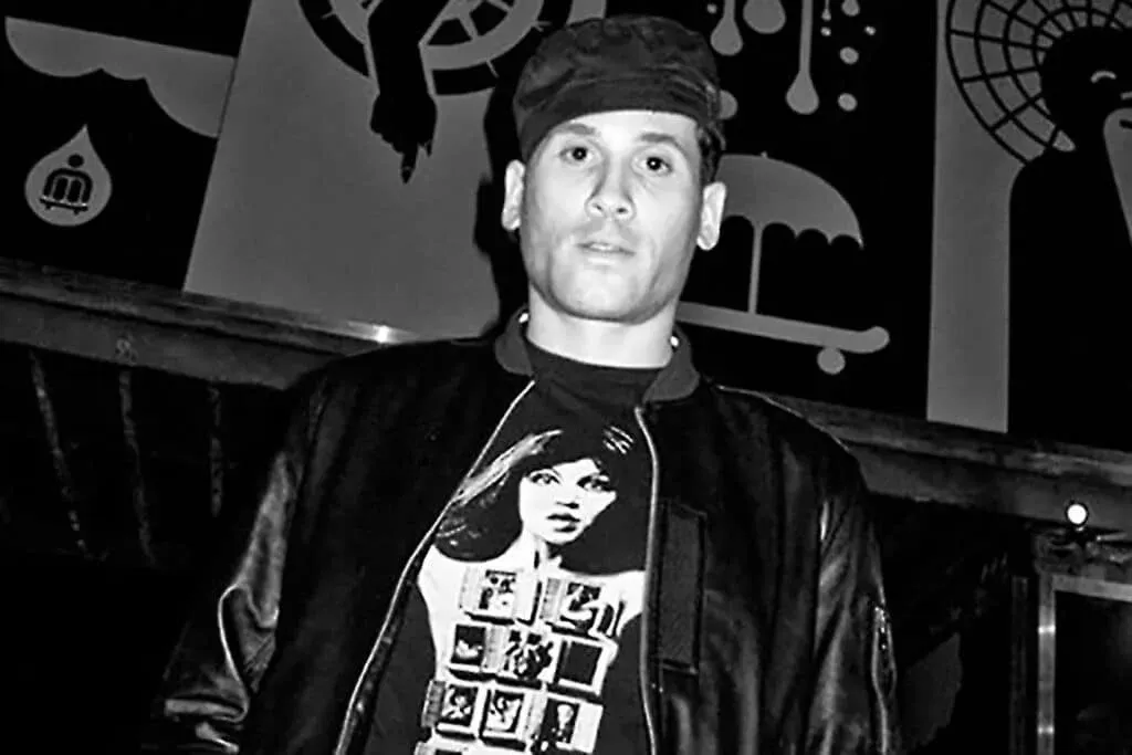 A person wearing a black jacket and a t-shirt with a graphic print, standing in front of a wall with abstract art designs in black and white.