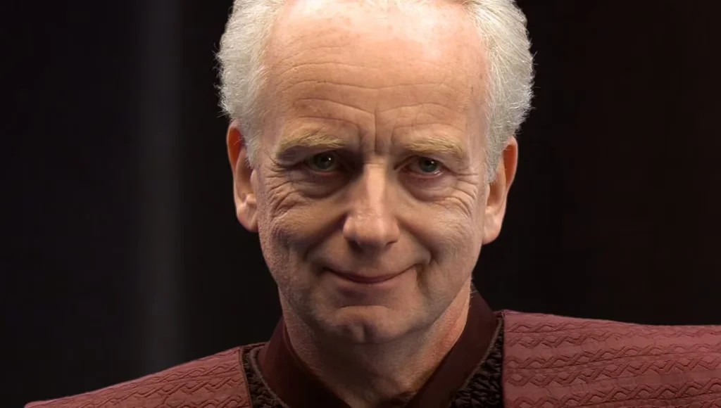 A man with light skin, white hair, and a slight smile wearing a textured burgundy outfit, against a dark background.