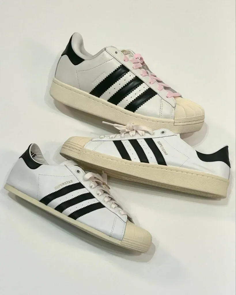 Three pairs of Adidas Superstar sneakers featuring the signature black stripes, white shell toes, and differing laces; some with traditional white laces and one with pink details, all arranged on a white background.