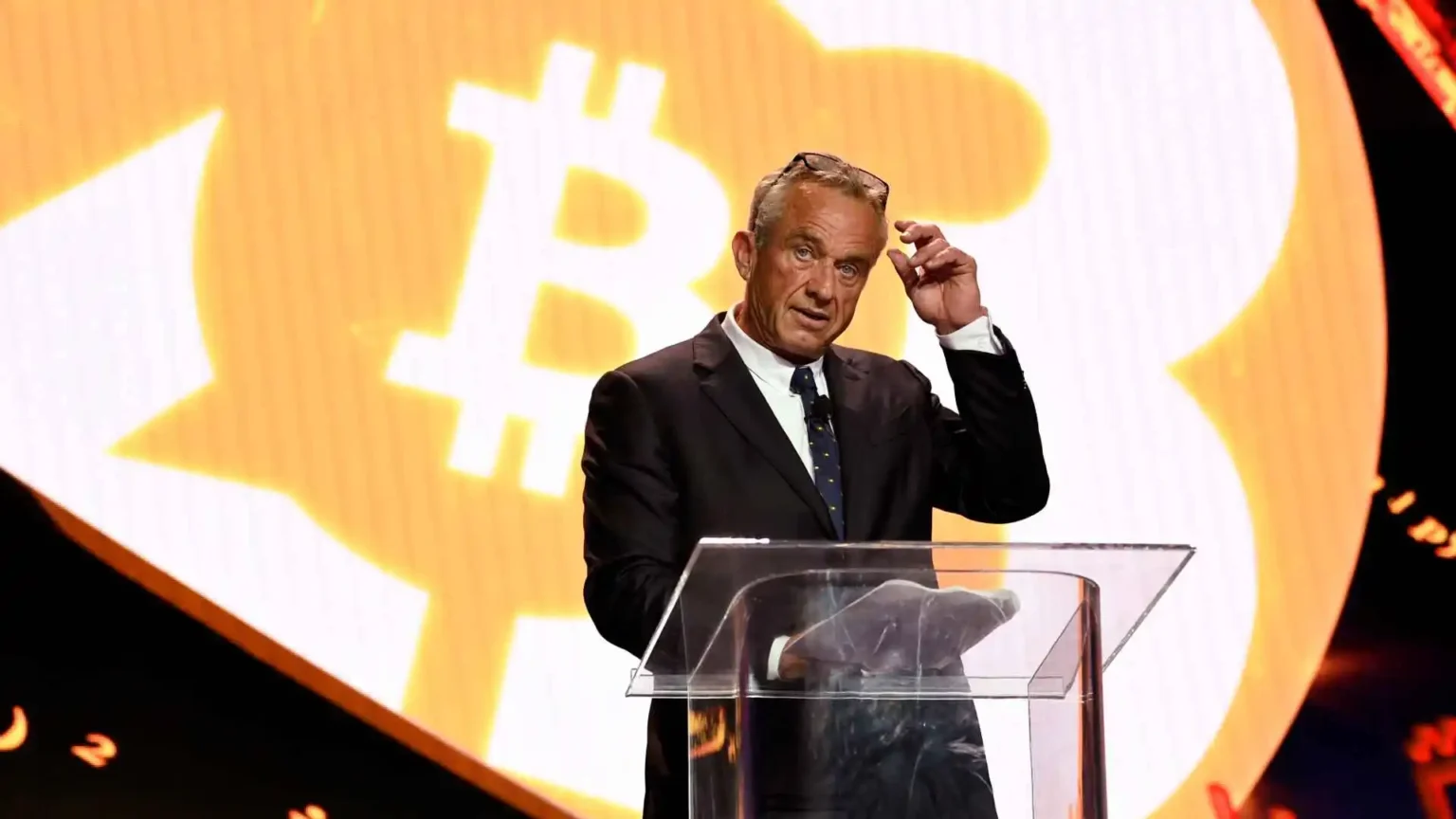 Man standing at a clear podium with a large Bitcoin symbol displayed on a screen in the background.