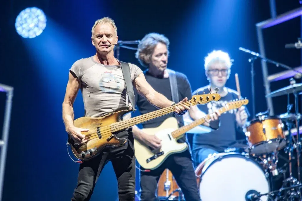 A musician playing a bass guitar on stage with a band; another guitarist and a drummer are visible in the background, all under bright stage lights.