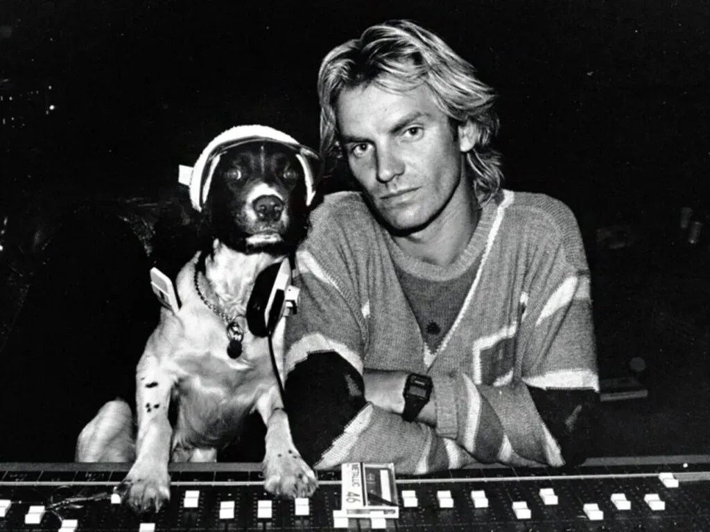 A black and white photo of a man with long hair sitting next to a dog wearing headphones in a recording studio, with both looking towards the camera.