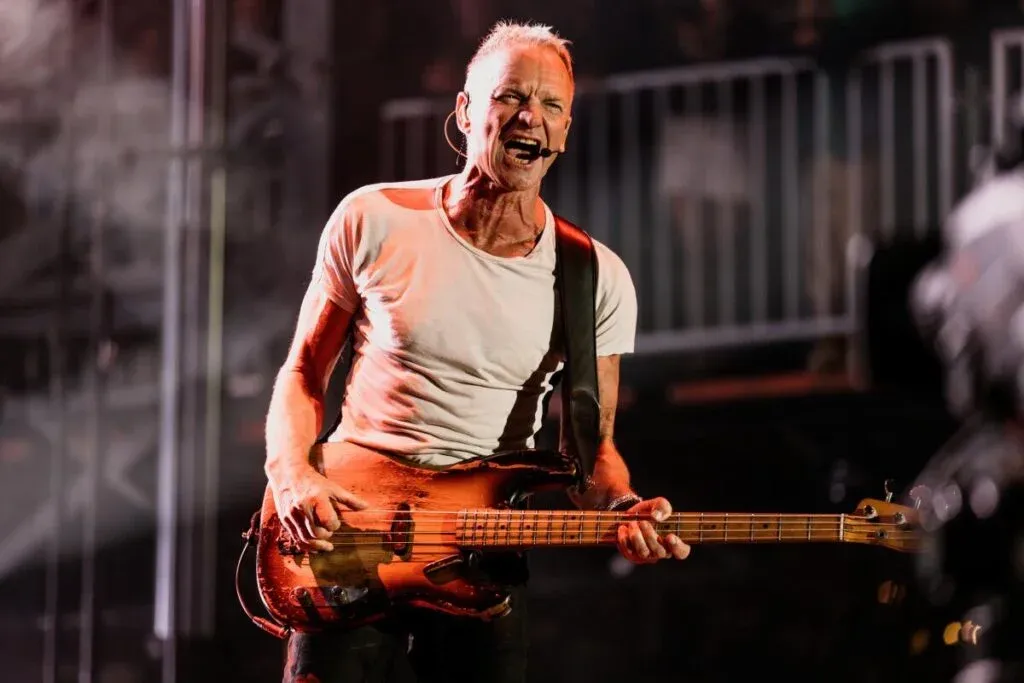 A musician passionately playing a bass guitar on stage, wearing a white t-shirt and using a headset microphone, with vibrant stage lighting accentuating the performance.