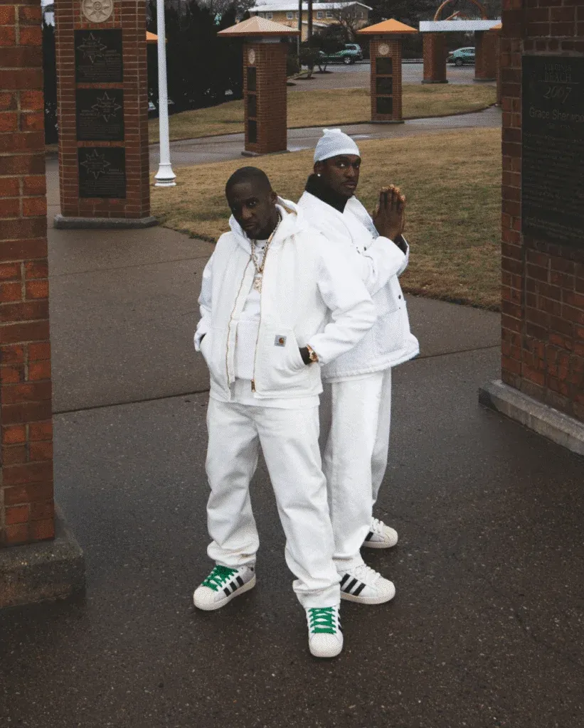 Two individuals stand in a park setting against brick structures, both dressed in all-white outfits with sneakers; one wears green-striped sneakers and the other black-striped sneakers.