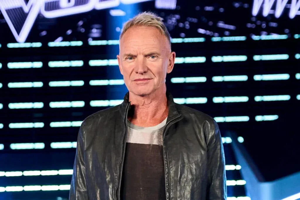 A person with short blonde hair wearing a leather jacket stands in front of an illuminated, digital background.