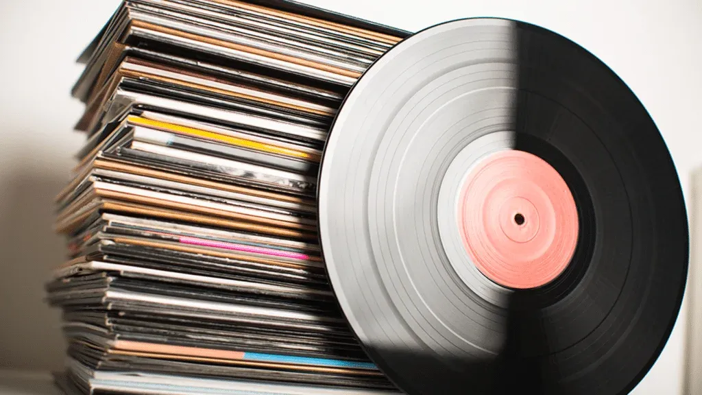 A large stack of vinyl record sleeves with a black vinyl record leaning against it, showing a partially pink label.