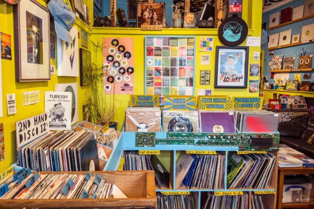 An eclectic record store with colorful walls displaying a variety of vinyl records in wooden and plastic bins. The walls are adorned with posters, album covers, and a variety of musical genres indicated by signs such as Techno / Electro, Disco House, and Tech House / Minimal. In the foreground, multiple crates are labeled with different musical styles and categories.