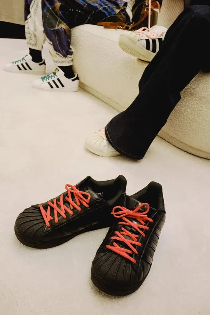 Adidas sneakers with bright red laces are on the floor, with a pair of feet wearing white sneakers with blue and pink laces visible in the background.