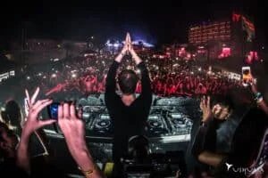 A DJ performs at an outdoor night event with a large crowd in front, illuminated by lights and camera flashes. The scene includes multiple DJ decks and visual effects, set against a backdrop of a brightly lit hotel building displaying the name 'Ushuaïa.' People hold up smartphones, capturing the moment.