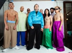 A group of men posing indoors, wearing brightly colored, oversized pants; some are topless, and others wear loose jackets and shirts. The background shows a plain wall and studio lighting equipment.