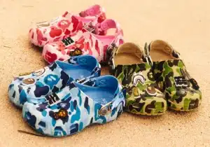 Three pairs of colorful, cartoon-themed Crocs shoes featuring camouflage patterns in pink, blue, and green, adorned with various decorative badges, displayed on a sandy surface.