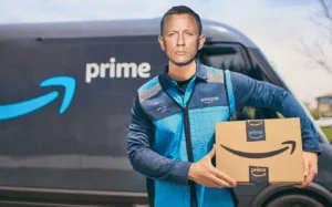 A person wearing an Amazon Prime delivery uniform holding a package in front of an Amazon delivery van.