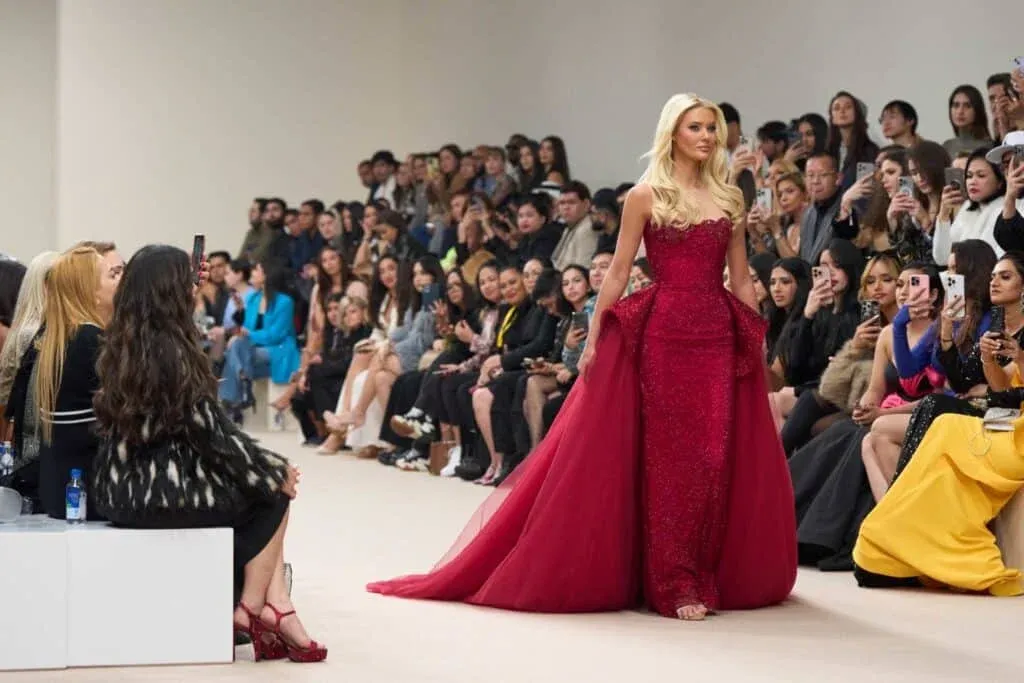 A model wearing an elegant red gown walks down a runway, with an audience seated on both sides, many capturing the moment with phones.
