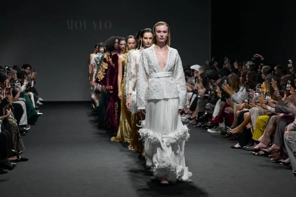 A group of models walking down the runway in elegant gowns during a fashion show, with a seated audience capturing photos on their phones on both sides.
