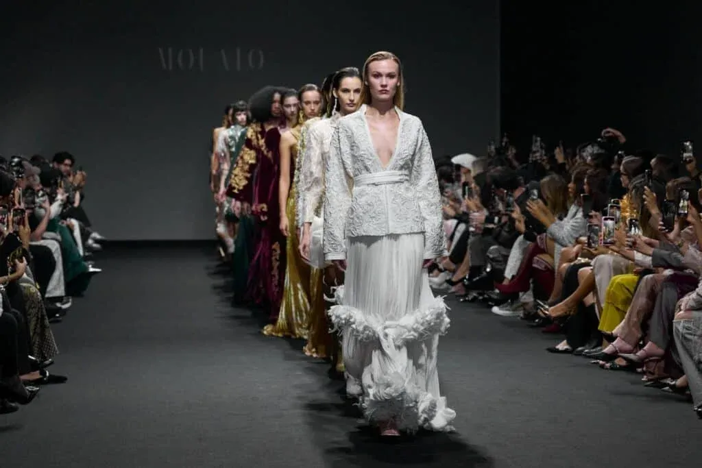 A group of models walking down the runway in elegant gowns during a fashion show, with a seated audience capturing photos on their phones on both sides.