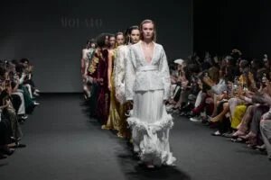 A group of models walking down the runway in elegant gowns during a fashion show, with a seated audience capturing photos on their phones on both sides.