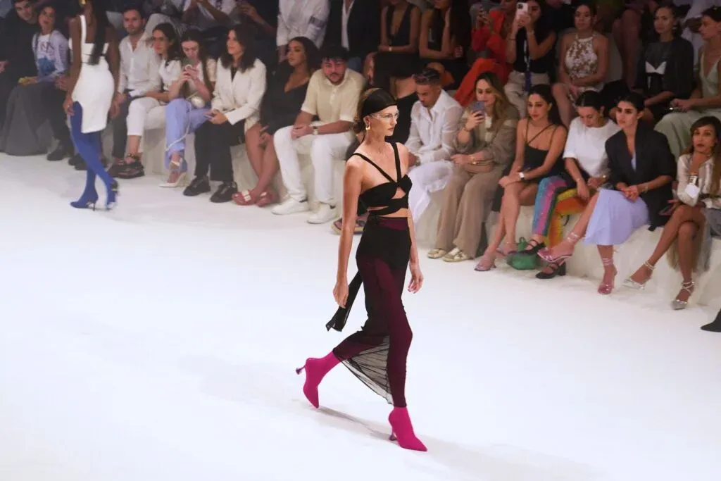 A fashion model walks the runway in a stylish black cut-out outfit with sheer panels and bright pink boots, surrounded by an attentive seated audience at a fashion show.