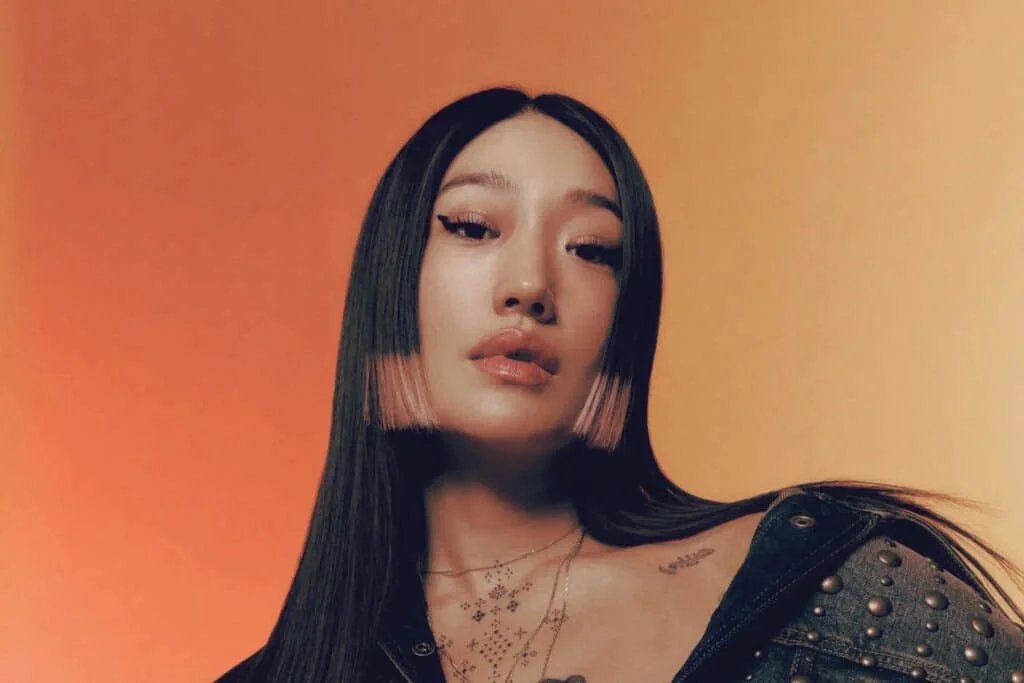 Portrait of a woman with long, sleek, black hair featuring pink tips, standing against a gradient orange background, wearing a dark studded jacket and layered necklaces with tattoos visible on her neck.