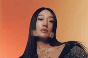 Portrait of a woman with long, sleek, black hair featuring pink tips, standing against a gradient orange background, wearing a dark studded jacket and layered necklaces with tattoos visible on her neck.