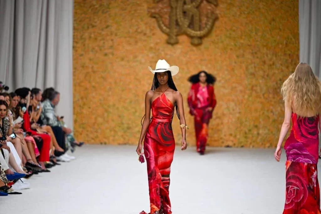 A model walking down a fashion runway wearing a red floral dress and a white cowboy hat, with an audience seated on the side and a blurred backdrop of orange and gold.