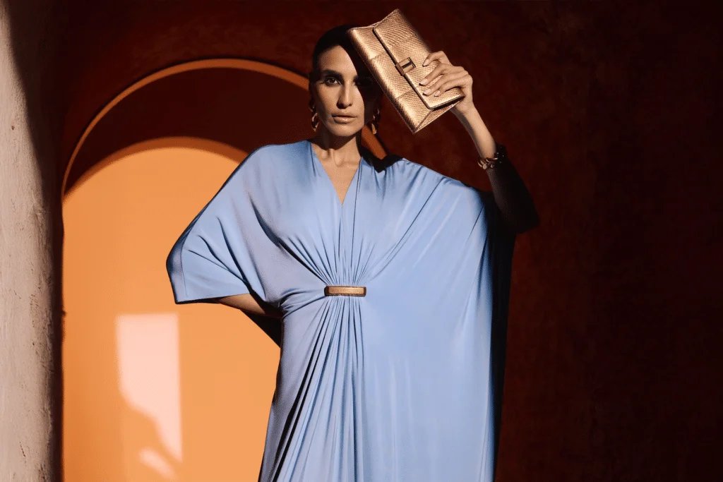 A person wearing a light blue draped dress holds a metallic gold clutch to their forehead, standing in a warmly lit arched doorway.