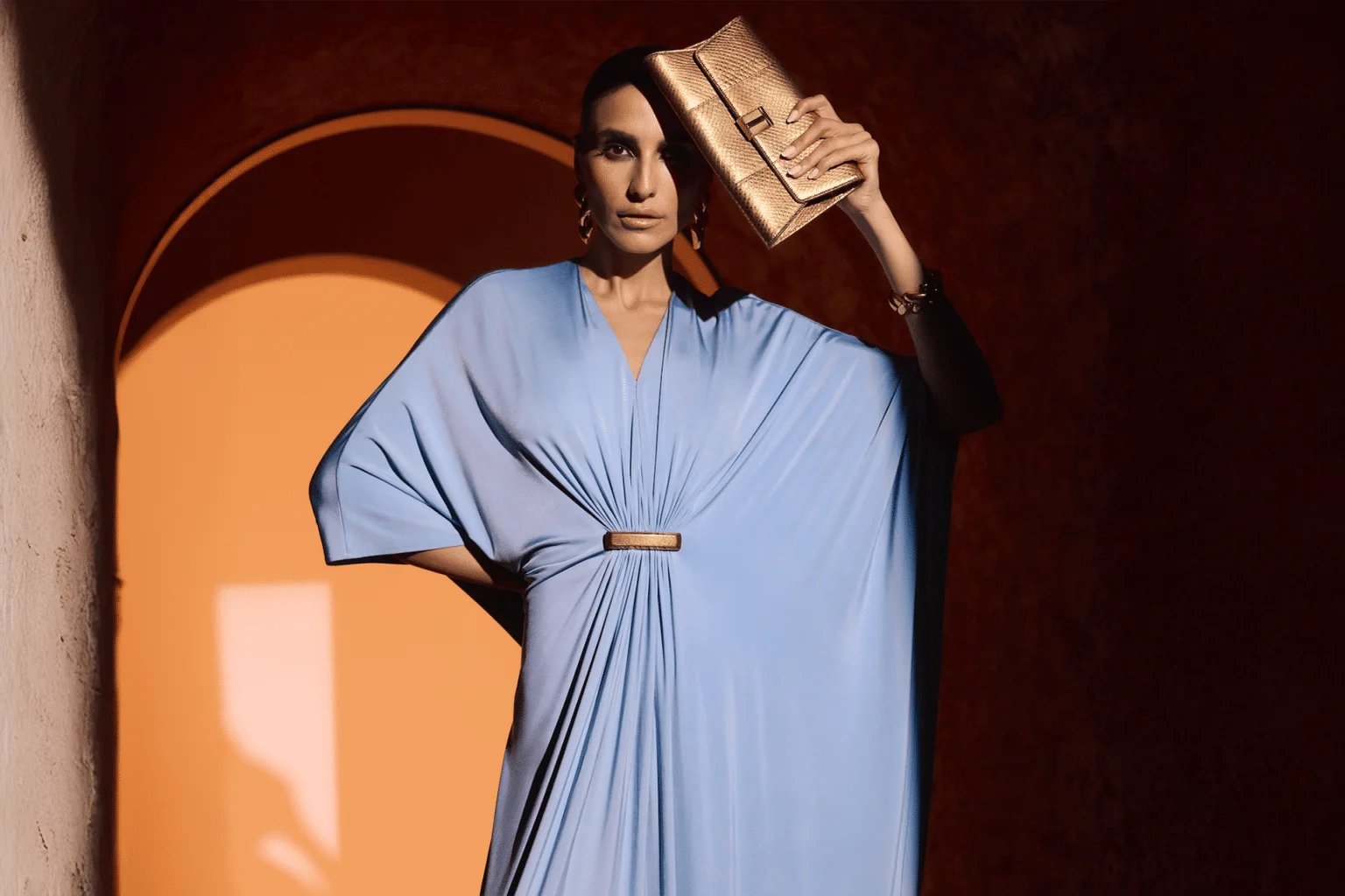 A person wearing a light blue draped dress holds a metallic gold clutch to their forehead, standing in a warmly lit arched doorway.