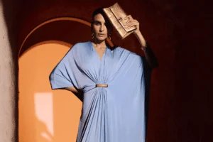 A person wearing a light blue draped dress holds a metallic gold clutch to their forehead, standing in a warmly lit arched doorway.