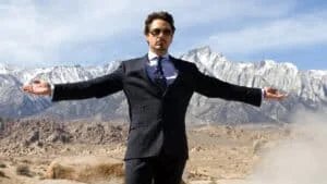 A man in a dark pinstripe suit, wearing sunglasses, stands with arms outstretched in front of a mountainous landscape with snow-capped peaks under a clear blue sky.