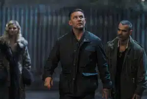 Three people standing in a dimly lit outdoor setting, with a man in a dark jacket looking upwards, flanked by a woman in a fur coat and another man in a hooded jacket.