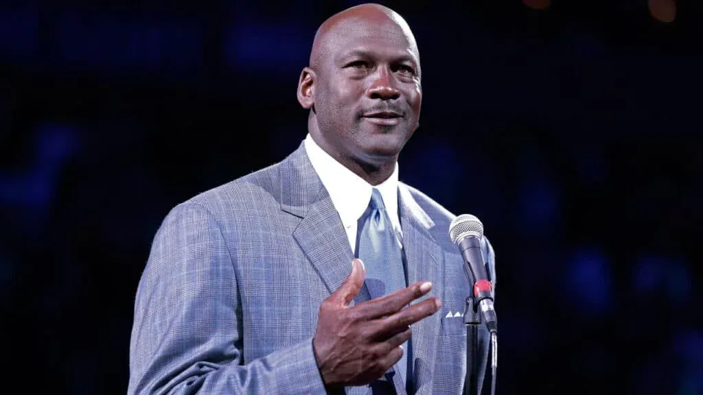 A man in a suit speaking into a microphone with a dark background.