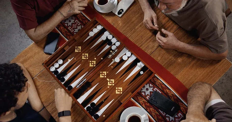 Three people sitting around a table playing a game of backgammon, with coffee cups and smartphones nearby.