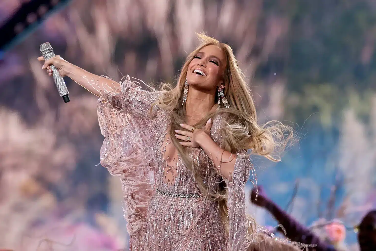 A singer with long hair, wearing a sparkling pink dress, joyfully performs on stage while holding a microphone and smiling widely.
