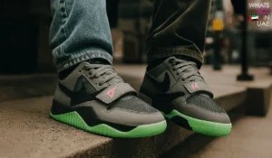 A close-up of a pair of sneakers featuring a dark grey upper with black accents and vivid green soles, worn by two people standing on an outdoor step. The shoes display unique design elements including a strap with a bright pink symbol.