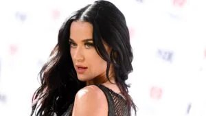 A woman with long, wavy dark hair and striking makeup is wearing a black, textured sleeveless top, posing against a blurred white background.