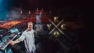 A DJ smiling in front of a massive crowd at a concert venue, with electronic equipment and keyboards around him. The letters 'K', 'Y', 'G', and 'O' are prominently displayed, intersecting in a cross pattern over the scene.
