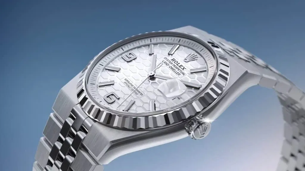 A luxury wristwatch with a patterned white dial, silver bezel, and metal bracelet, featuring hour markers and a date window.