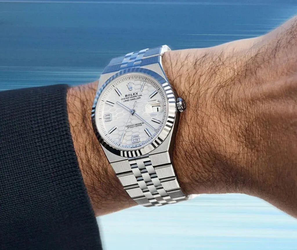 A silver wristwatch with a white textured dial and stainless steel bracelet on a person's arm against a blurred blue background.