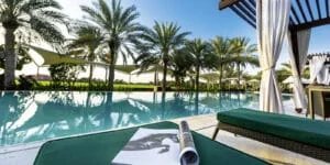 A tranquil poolside setting featuring a clear swimming pool bordered by palm trees and shaded areas. In the foreground, there is a green lounge chair with an open magazine displaying a black-and-white image of a horse.