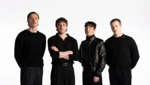 Four men standing side by side, all wearing black outfits, against a plain white background.