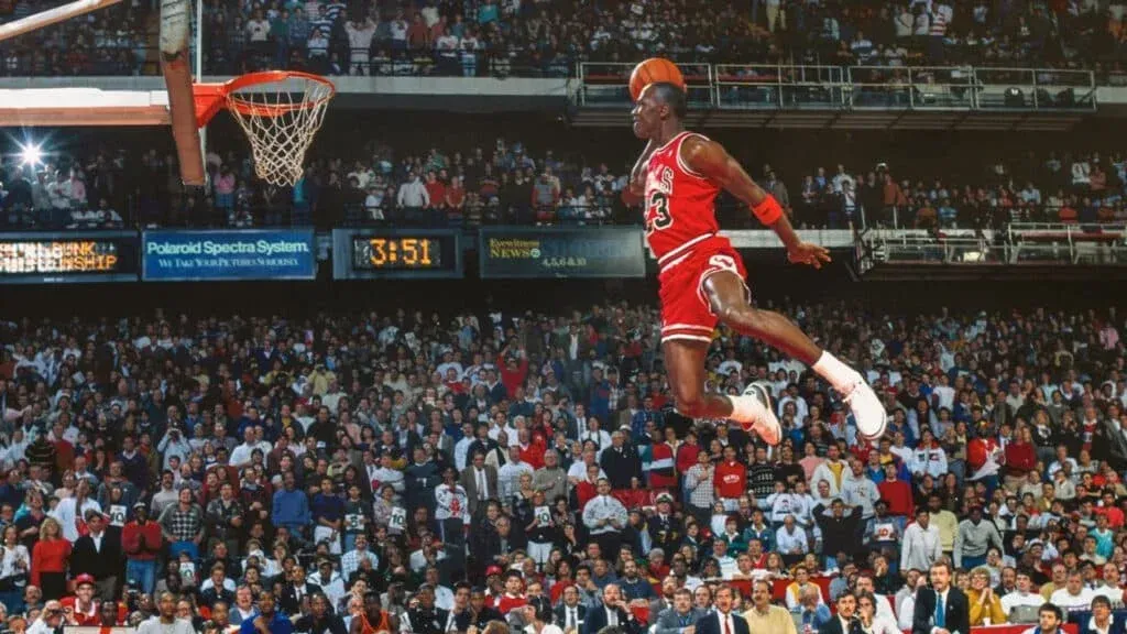 A basketball player in a red uniform is mid-air performing a dunk in front of an audience in a large stadium. The scoreboard and various advertisements are visible in the background.