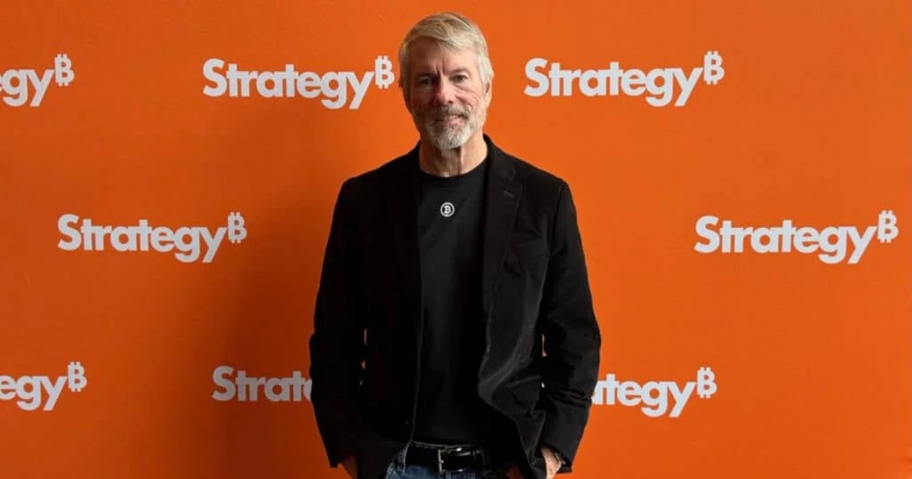 A man with a beard and mustache stands in front of an orange backdrop with repeating white text "Strategy B." He is wearing a black blazer over a black T-shirt featuring a Bitcoin logo, along with jeans.