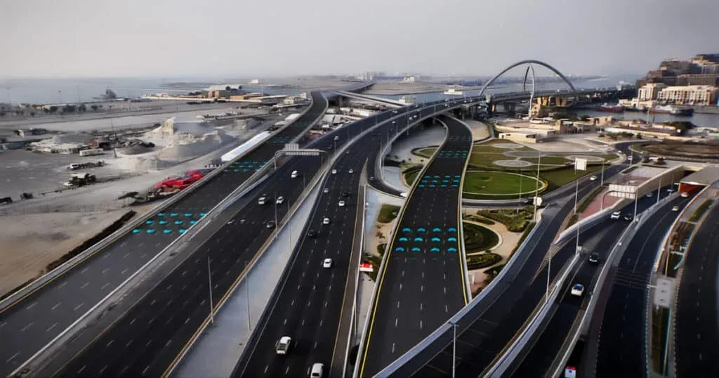 Aerial view of a multi-lane highway interchange with several vehicles, featuring a modern arch bridge and surrounded by landscaped areas and sparse industrial buildings in the background.