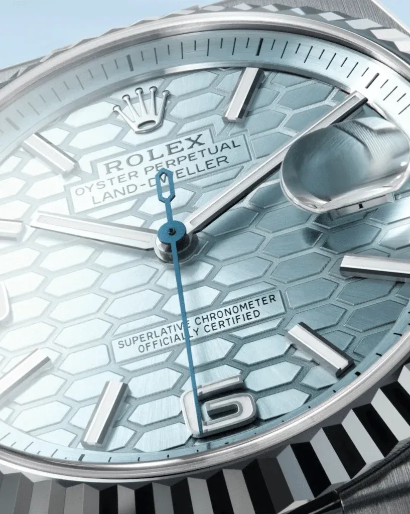 Close-up image of a luxury watch face with a light blue honeycomb pattern, featuring silver hour markers, a blue second hand, and a date magnifier over the number 16.
